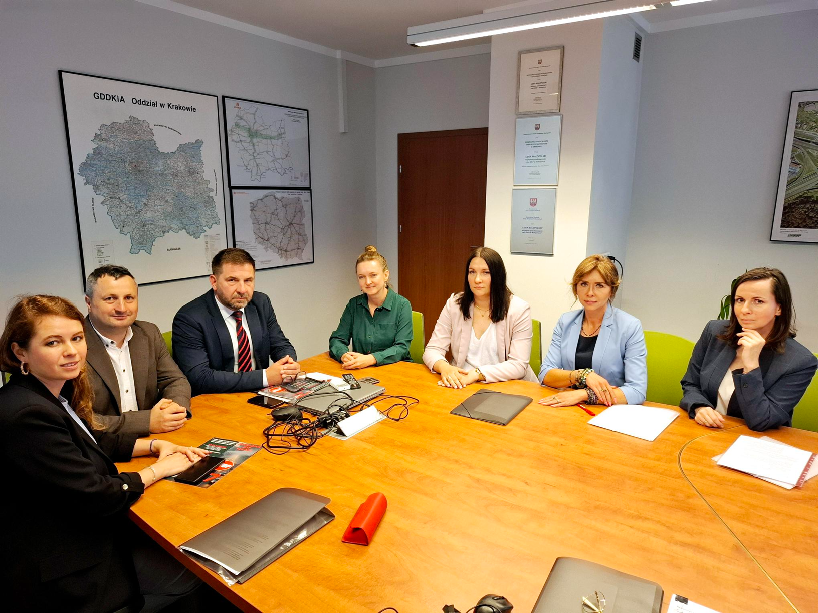 Siedem osób – pięć kobiet i dwóch mężczyzn – siedzi przy stole konferencyjnym w sali biurowej. W tle widoczne są mapy i certyfikaty na ścianach. Na stole leżą dokumenty, laptopy i przewody. Wszyscy uczestnicy patrzą w stronę obiektywu. Atmosfera spotkania jest formalna.